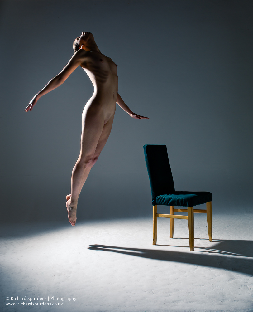 Dance Photography - Dance Photographer - colour image of dancer doing a small jump with a chair in the near back ground with her hand held out to the side she looks if she is levataing around the chair