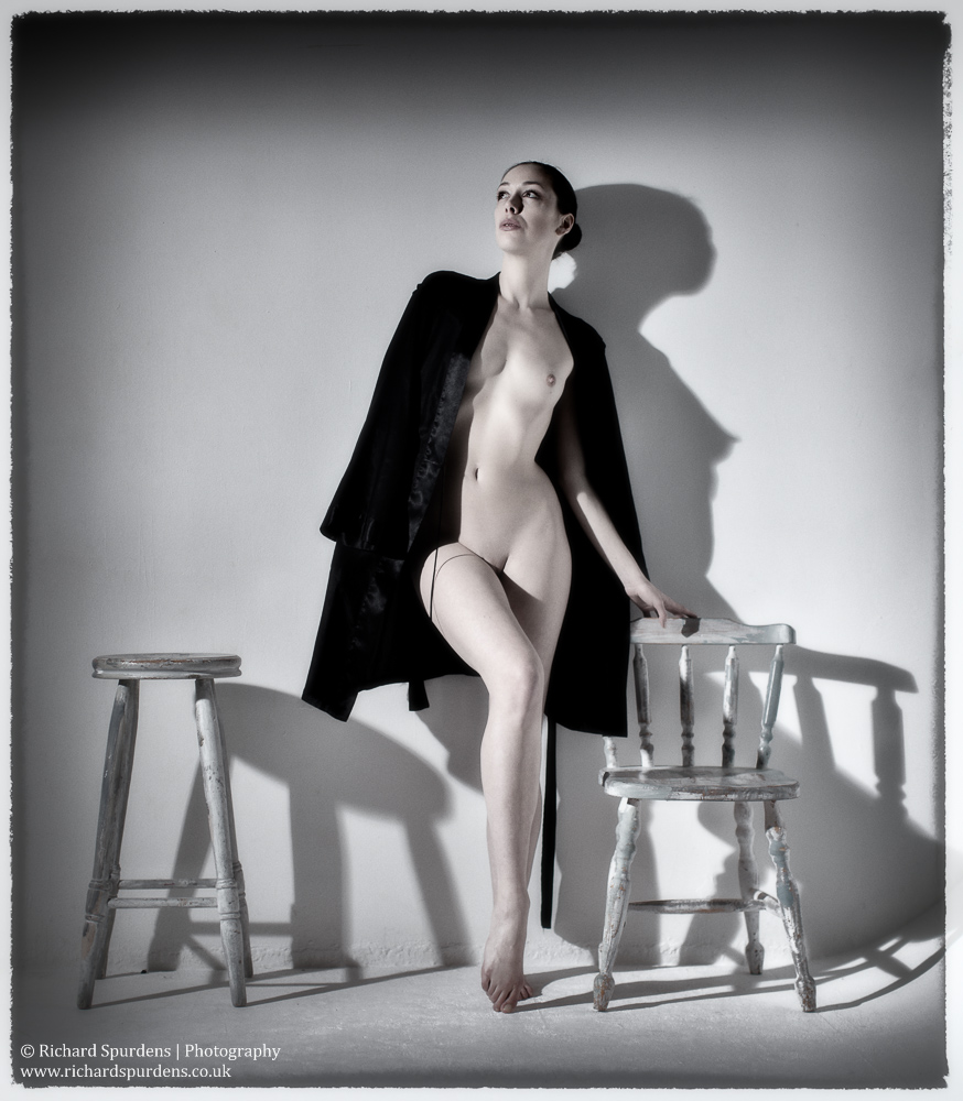 Fine Art Nude Photography - Fine Art Nude Photographer - muted colour image of dancer leaning against a wall with hard shadows of her figure and the chair and stool she is stood between