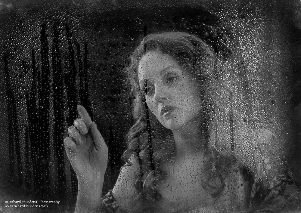 portrait photography - portrait photographer - monochrome image of a model standing behind a window with rain drops runing down the outside. she is reaching her finger to trace a rain drop down the window - rainy day reflections