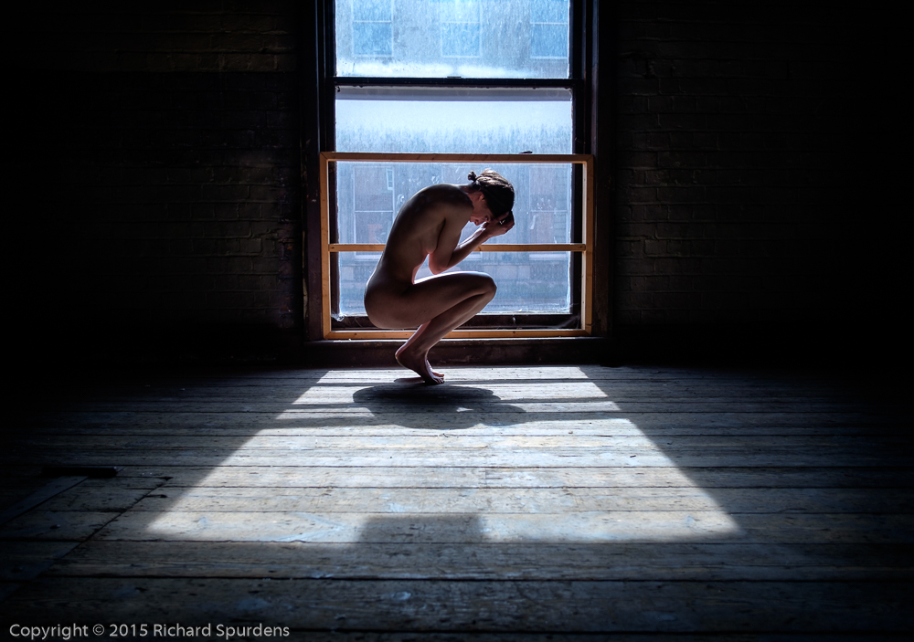 Artistic nude photographer - artistic nude photography - model crouching in the window light on balls of her feet and holding her head in her hands exposed for the window light so models figure is in shadow