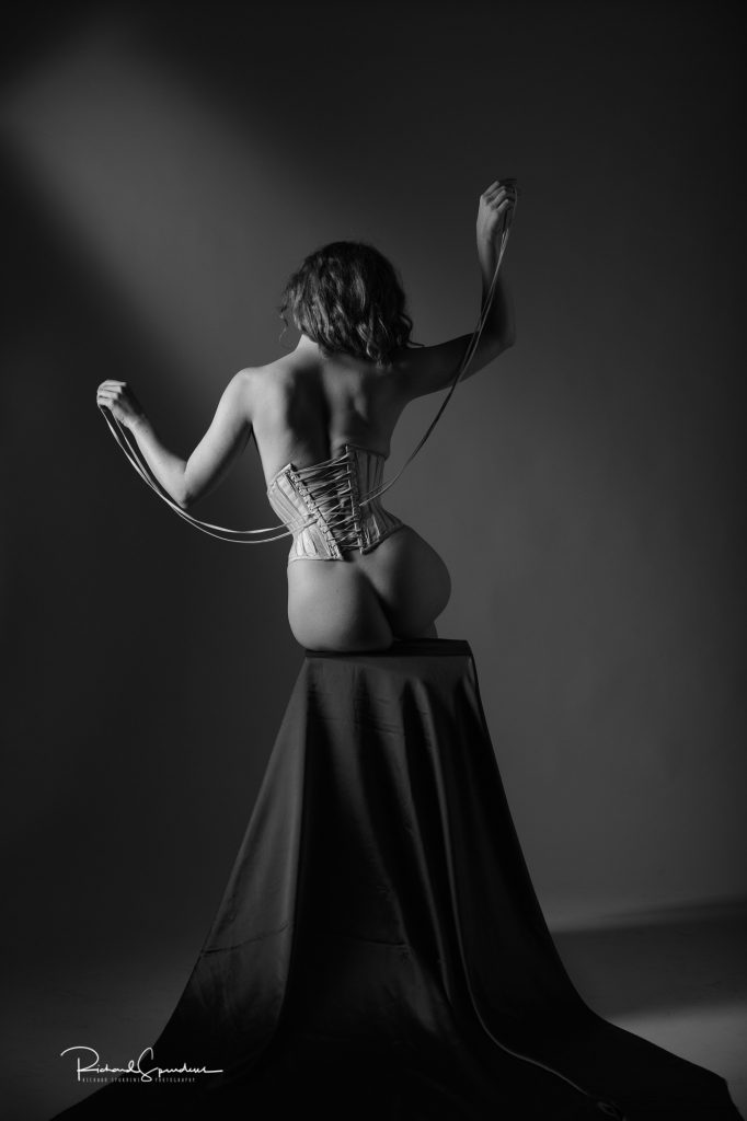 Artistic nude photographer - artistic nude photography - monochrome image of model seated on a tall seat she is wearing a laced corset and is holding the laces out to the side to make a pleasing shape with the laces