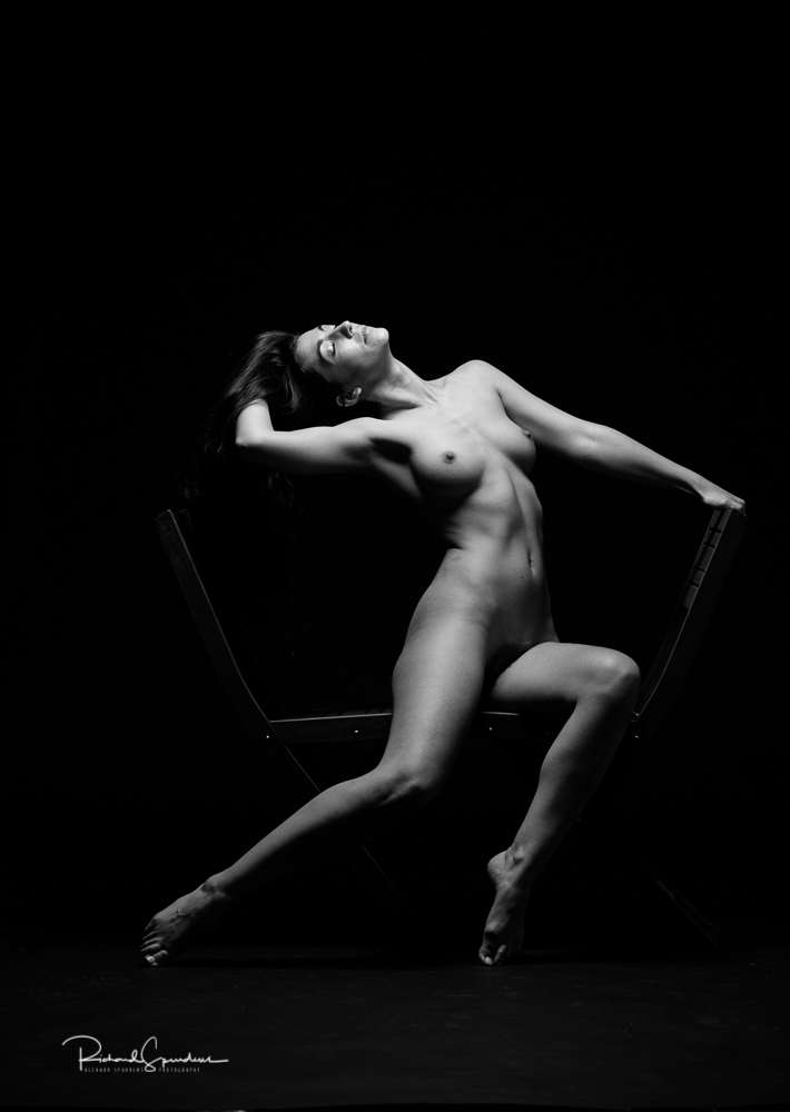 Artistic nude photographer - artistic nude photography - single light monchrome image of model make shapes and angles on the edge of two wooden chairs the light is playing along to edges of her legs and arms to make strong highlights on those areas and deep shadows in on the rest of her figure