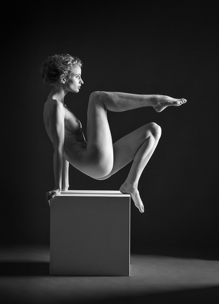 artistic nude photographer - artistic nude photography - monochrome image the model in using a white posing box she pushing up with her arms at the back of the box and her left foot to lift herself off the box her other leg is lifted and pointed at ninty degress towards the light. which is a single side light to the right hand side of the model.