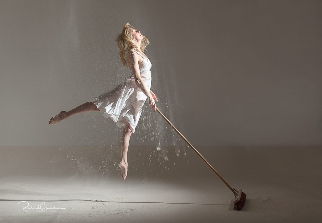 dance photographer - dance photography - colour image of the model wearing a white dress and leaping in the air with one leg pointing vertically down and the other out behind her she is hold a large sweeping brush in her right hand floor has been drop on her from above to make it look like she is sweep up magic dust