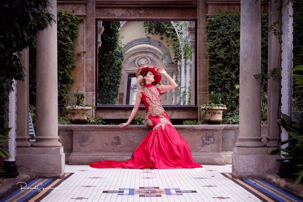 fashion photographer - fashion photography - colour image of model wearing a red and gold ball gown with makeing red hat she is seated on a stone plinth in the garden room- shot on location at sandon hall, Dress Designer: Katie Newsam, Styling Jen Brook, Walter's Wardrobe Productions