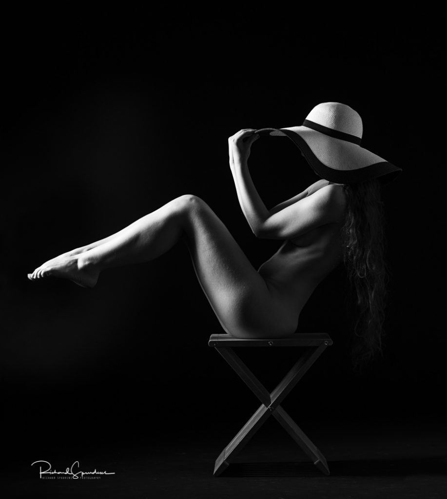 Fine Art Nude Photography - Fine Art Nude Photographer - monochrome image of artistic nude model sitting in profile on a small wooden stooll and her legs are raised forward to form an angle she is wearing a white hat and her arms are both reaching up to hold the hats brim. lit with a single gridded rectangular soft box