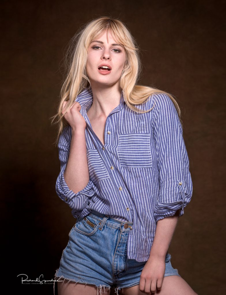 fashion photographer - fashion photography - colour image from a fashion shoot with model wearing croped demin jeans and a striped shirt she is stood against a dark brown back ground with one arm hold the shirt coller and the other resting on the opposite leg