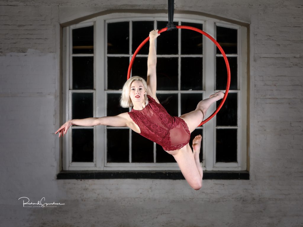Aerial Arts photographer - Aerial hoop photographer - Aerial Arts photography - colour image of aerialist on a red hoop and wearing a red leotard holding an aerial pose using hand one hand and inner leg