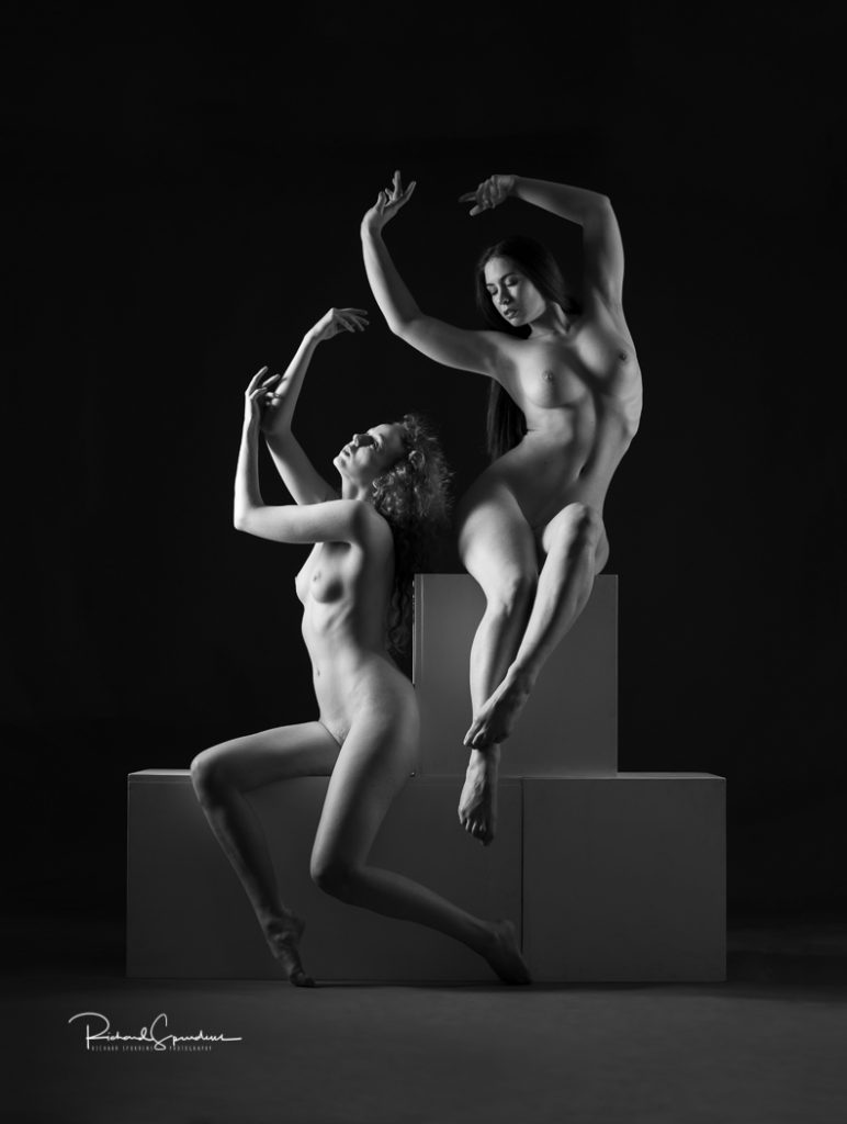 artistic nude photographer - artistic nude photography - monochrome image of two artistic nude seated on white posing blocks one is sat sideways with her legs bent at the knees to make a triangle shape, the other how is slightly higher on the blocks at is arching her figure to the side with her legs crossed at her feet, they are both reaching up towards the light that is coming in from the lhs,