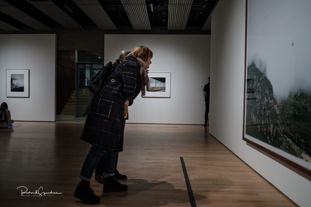 travel photography - travel photograher - colour image from a visit to the hayward gallery focusing on the visitors viewing the Gursky images two visitors lean forward towards a large image of a landscape the angle make the image as the rest of the Image is squares and vertical lines