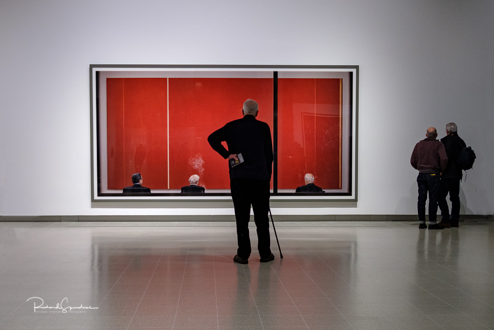 travel photography - travel photograher - colour image from a visit to the hayward gallery focusing on the visitors viewing the Gursky images a man with a walking stick stand and views the fashion shot of the four head of state from the back sat in front of a red wall
