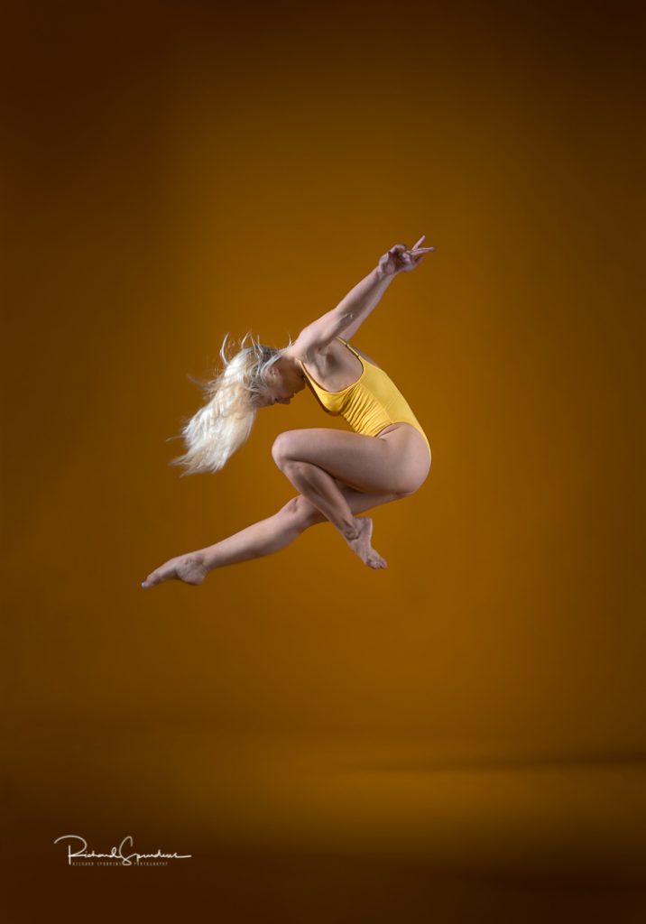 dance photographer - dance photography - colour image of dancer leaping up with one leg pointed out straight and the other bent at the knee to make a right angle , he arms are reaching out backward , she is wearing a yellow leotard and is working against a yellow background shot in profile