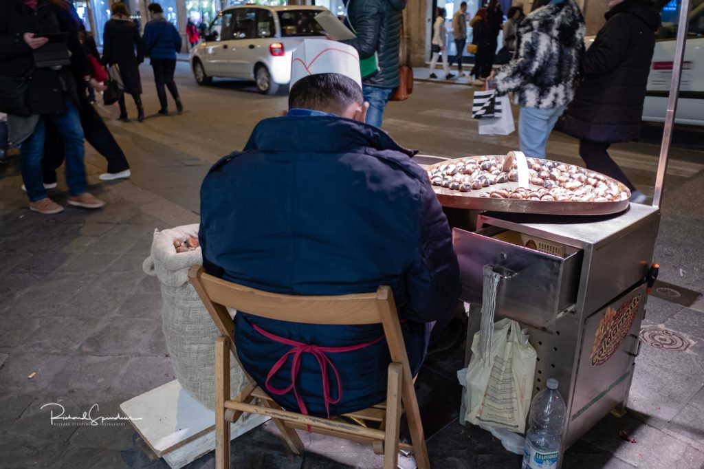 travel photography - travel photograher - colour image from a visit to rome shot from behind a street food seller he is selling warm chestnuts and is illuminated by his stands lights in the back ground shopper walk be with there warm coats and shopping bags