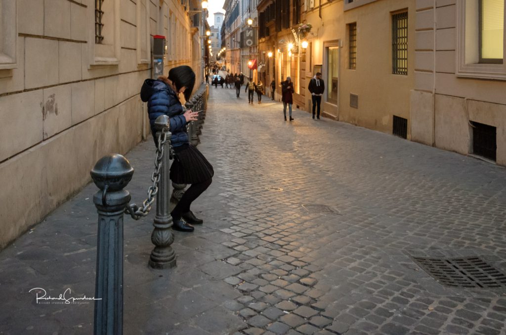 travel photography - travel photograher - colour image from a visit to rome this one a street image spotted on a side street a lady sitting on a chainlink fence lost in here phone (february images)