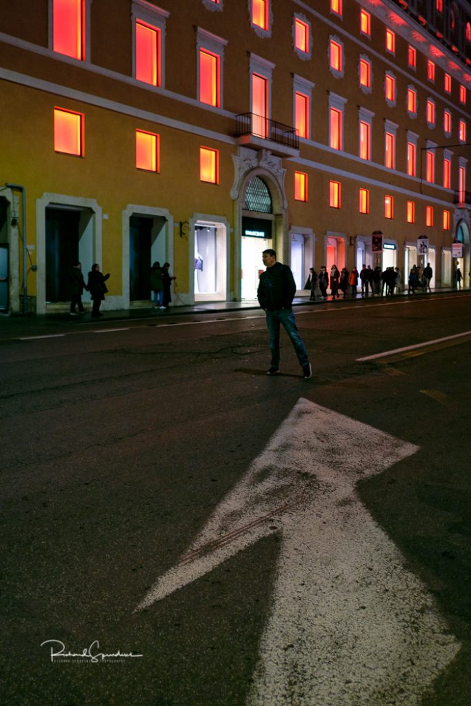 travel photography - travel photographer - image showing a large road lane direction arrow pointing to figure looking down the road against a large build where all the windows have an orange glow coming from them (photographing rome at night)