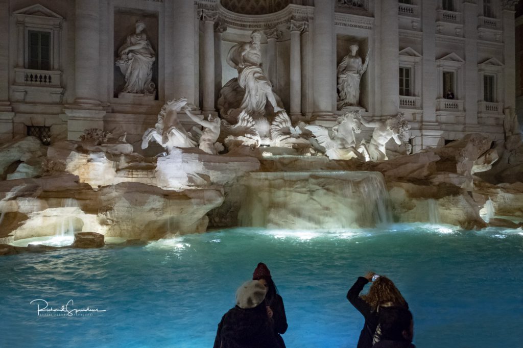 travel photography - travel photograher - colour image from a visit to rome this one from the trevi fountain square shot around 20:00 in the bottom are three tourist one is taking a phone picture the other two are looking at the results of the self they took in the back ground is the fountain lit by the flood lights