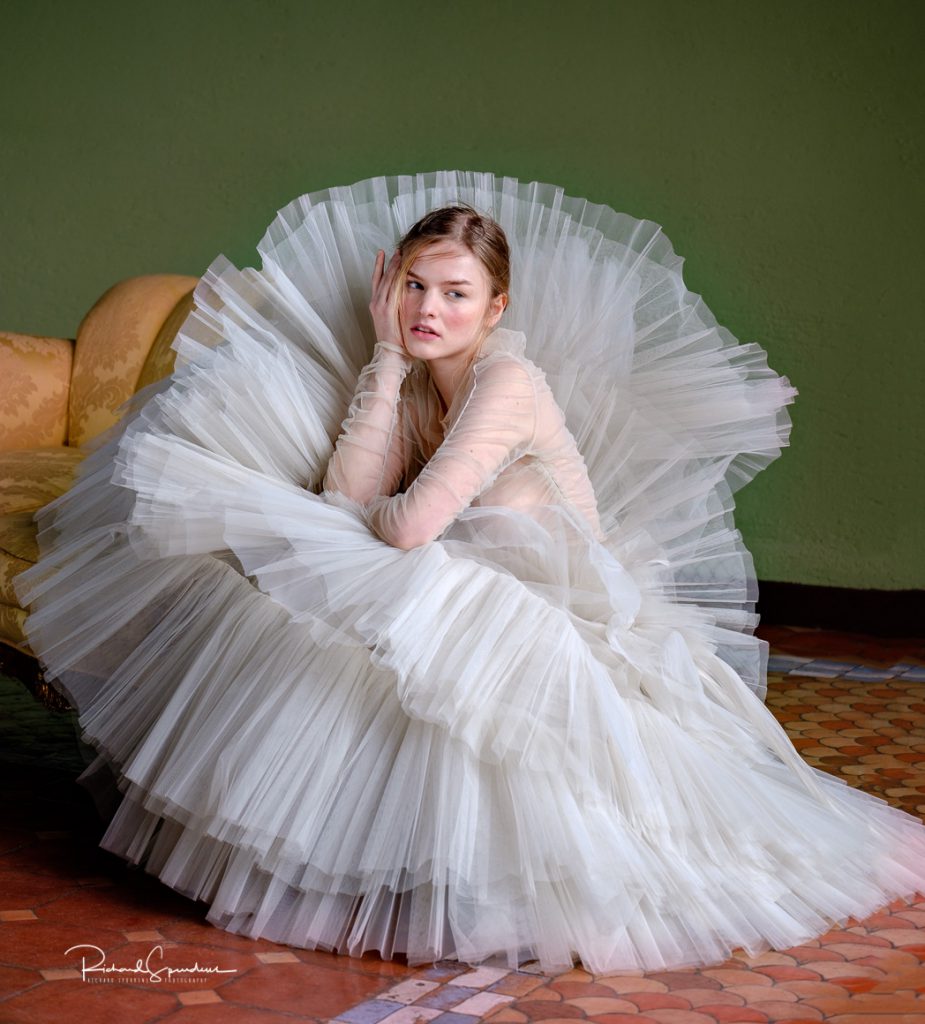 fashion photographer - fashion photography - colour image from a fashion shoot at villa giovanelli near rome - the model is crouching down surrounded by hte crimpeleen skirts of the white dress she is look towards the window ltight