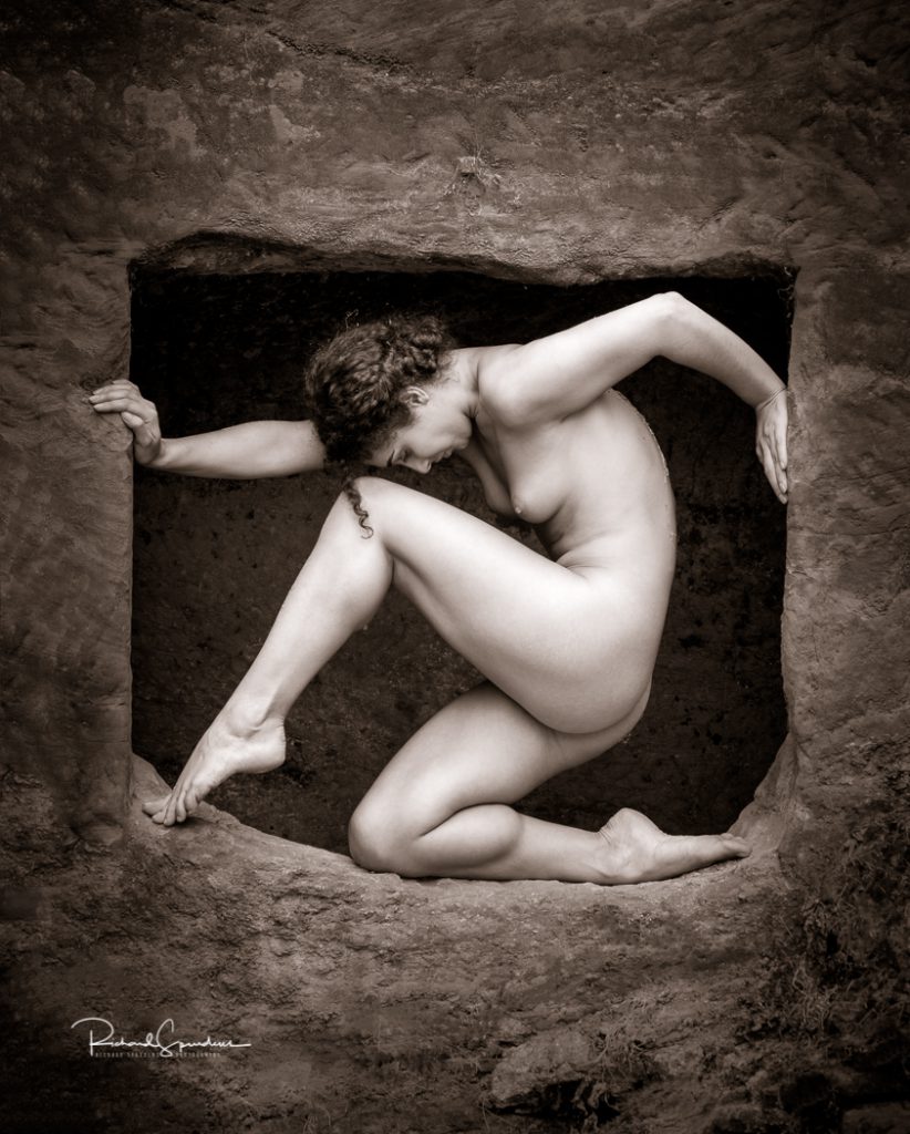 artistic nude photographer - artistic nude photography - a tonned monochrome image artistic nude model Micshkan she is posing in a rock opening, her arms are pushing at the sides and she is nearing on one leg and the other is pointed into the opposite corner. shot on location Guys Cliffe.