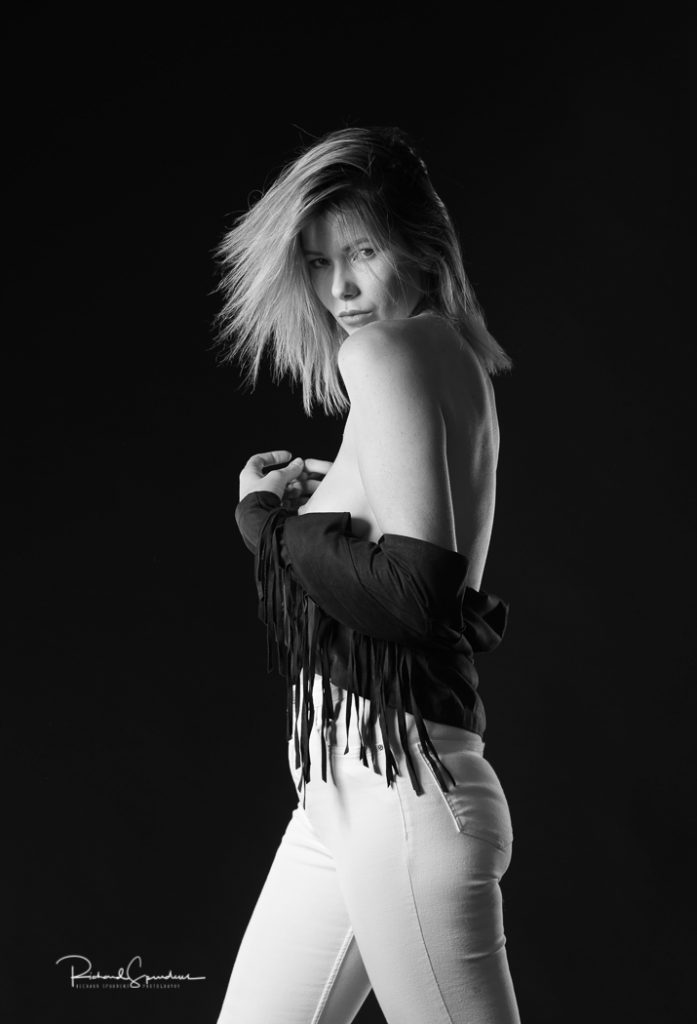 fashion photographer - fashion photography - monochrome image the model wearing white jeans and a suede jacket off her shoulder and across her middle she is turned side ways with her head turned to look back at the camera