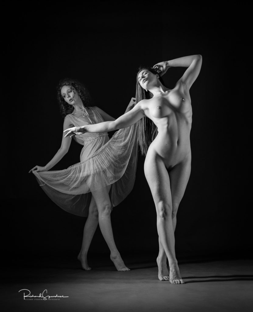 Artistic nude photographer - artistic nude photography - monochrome image of artistic nude models elle beth and Gem working as a duo pair with elle B on tip toes and gem behind wearing a floaty dress the side lighting is highlighting elle beth figure with gem slight further back with the light picking up the edge of her dress