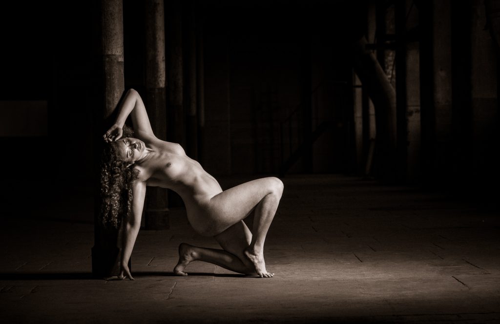 artistic nude photographer - artistic nude photography - a toned moody monochrome image using a single side light to illuminate the model fiqure as she creates a figure shape against the steel roof support post she is kneeing on her left leg and her right leg is bent at the knee with her foot on tip toe she is leaning back on a straight right arm and her left arm is bent at the elbow and rest on her head