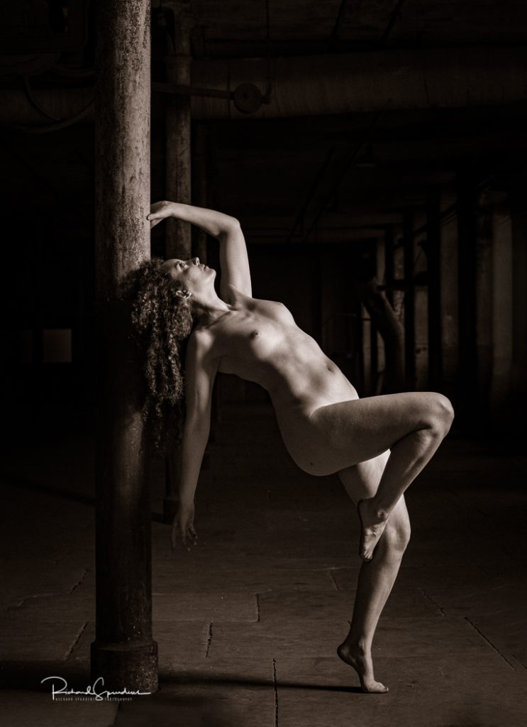 Artistic nude photographer - artistic nude photography - a toned monochrome image using a single side light to illuminate the model fiqure she is leaning back against a steel roof support her right leg is on tip toe and her left leg is bent at the knee she is touch to post with her head and left hand