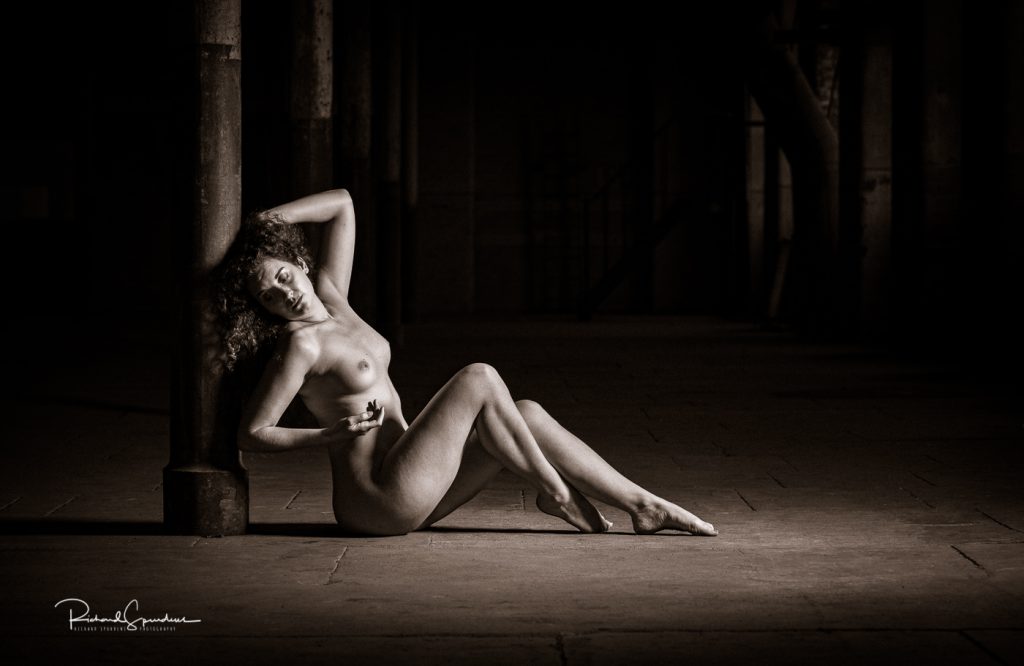 artistic nude photographer - artistic nude photography - monochrome image using a single side light to illuminate the model fiqure she is seated side on to the camera and her legs are bent at the knees and pointing out towards the light. her head is rest on the steel support post and her head is turned towards the camera and her arms are forming triangles on at her side and the other up to her head