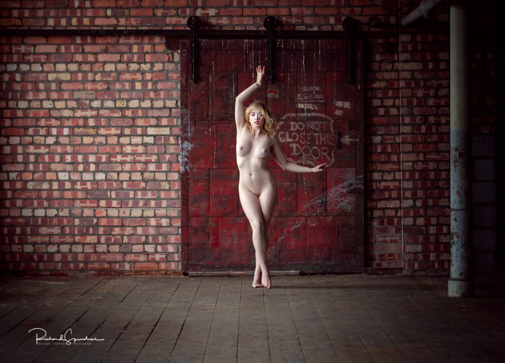 artistic nude model nicole rayner posing against a red steel door in an old mill location
