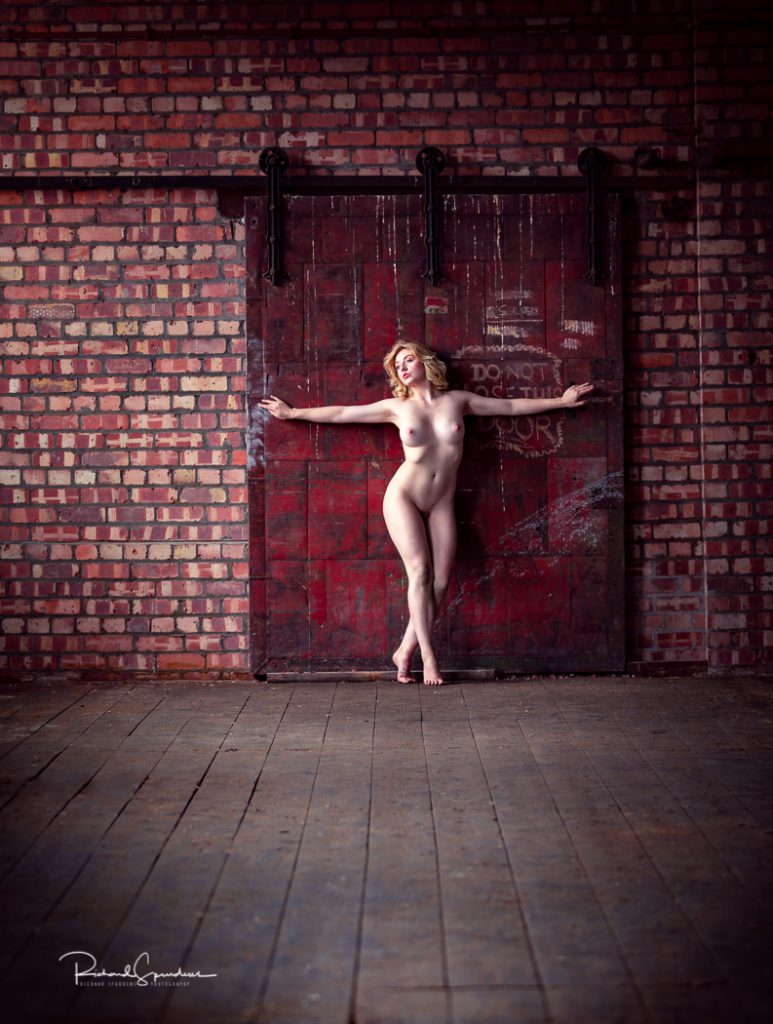 artistic nude model nicole rayner posing against a red steel door in an old mill location