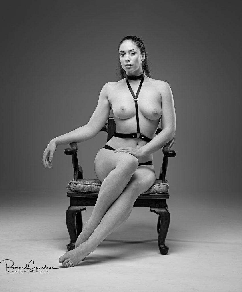 artistic nude photography - artistic nude photographer - monochrome image of model elle beth wearing a simple black leather harness and posing towards the camera with a relaxed look with her arm resting on the arm of the chair and on her forward knee