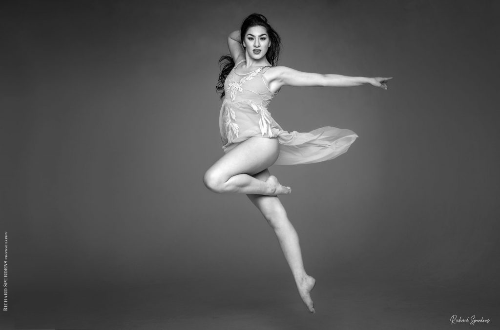 Dance Photographer - Dance Photography - monochrome image of dancer Elesha in mid air with front leg bent at ninty degrees and the back leg pointing down her left arm is pointing straight to the right of the image