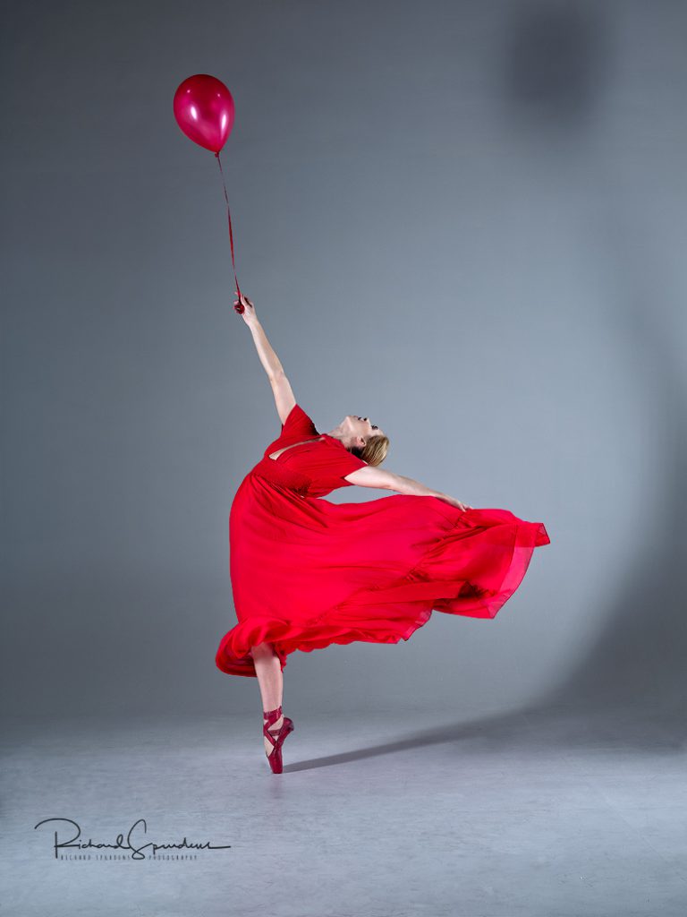 Dance Photographer - Dance photography - Image showing a dancer on pointe, wearing a red dress and holding a red balloon is it seems to pull her to the sky