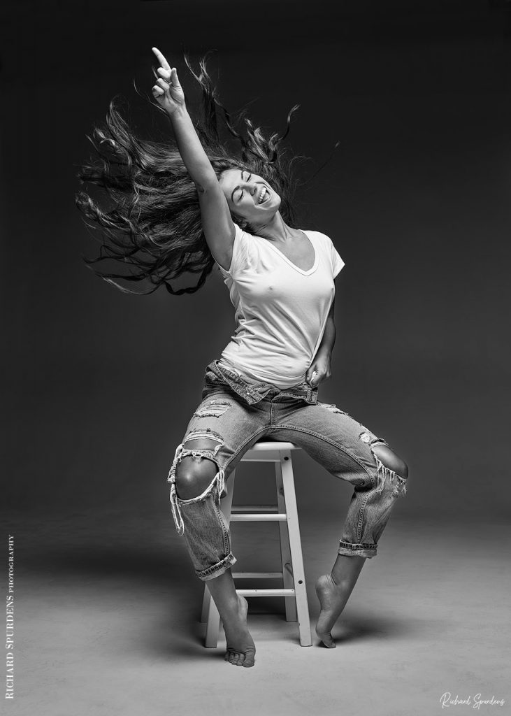 Fashion Photography - Fashion Photographer - monochrome image of natalia wearing torn jeans seated on a stool reach and pointing her right arm out and up to the ceiling with her hair flowing out backwards