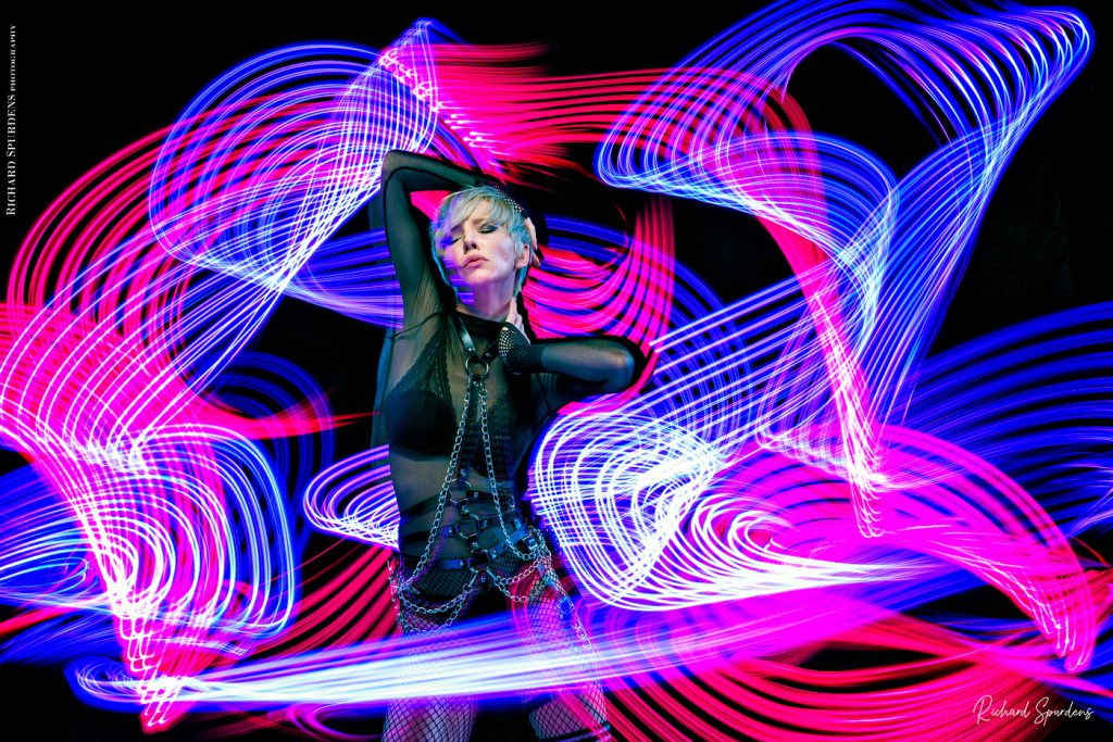 Light painting Photography - Fashion Photographer -- colour image of model wearing black underware and fetish chains and straps with lighting swirls around the model lighting swirls around the model using blues purple and white lights wands arouind the model