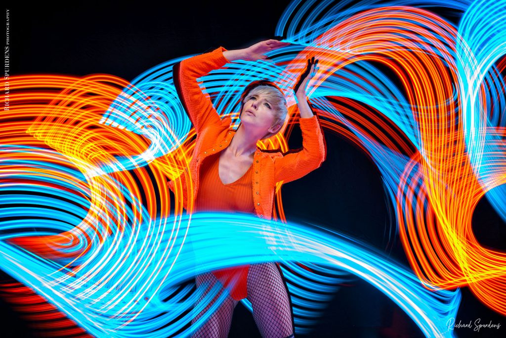 Light painting Photography - Fashion Photographer - colour image of model wearing orange jackets and body lighting swirls around the model using blues and orange lights wands around the model